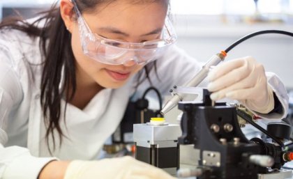 a woman in safety glasses looks closely at a yellow disk on a stand